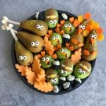A plate of fruit and vegetable bites arranged as cute Thanksgiving characters, including pears and kiwis decorated as turkeys and pilgrims with candy eyes, carrot details, and melon leaves for a fun touch of health.