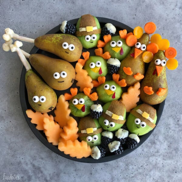 A plate of fruit and vegetable bites arranged as cute Thanksgiving characters, including pears and kiwis decorated as turkeys and pilgrims with candy eyes, carrot details, and melon leaves for a fun touch of health.