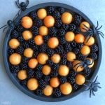 A round black platter filled with blackberries and orange melon balls, decorated with four large plastic black spiders around the edge, creates a Halloween-themed fruit arrangement perfect for healthy bites and festive food recipes.