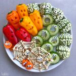 A plate of Halloween-themed veggie bites: mini peppers carved like jack-o’-lanterns, cucumber slices with candy eyes, broccoli “brains” topped with mayonnaise, and carrot spiders on pretzel webs—spooky fun for health-conscious food lovers.