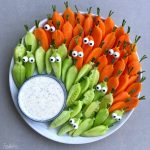 A plate of sliced carrots and cucumbers arranged upright with edible googly eyes and chive stems for a fun, cartoon veggie bite—served with a bowl of creamy dip on the side for a healthy, playful dish.