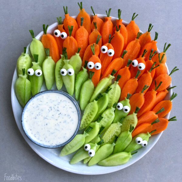 A plate of sliced carrots and cucumbers arranged upright with edible googly eyes and chive stems for a fun, cartoon veggie bite—served with a bowl of creamy dip on the side for a healthy, playful dish.