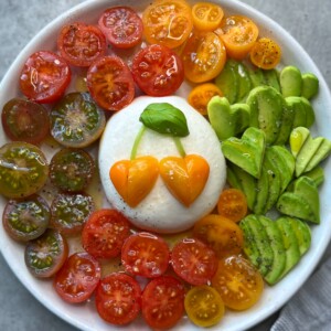 A bowl with colorful halved cherry tomatoes and sliced avocado arranged around a ball of mozzarella, topped with orange tomato halves and basil leaves for a cherry design—this vibrant tomato burrata salad is drizzled with olive oil and pepper.