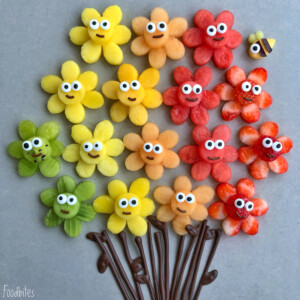 Colorful flower-shaped fruit bites with candy eyes are arranged in rows like a bouquet with chocolate stems. A small bee made from fruit rests near the top right flower, making these playful treats perfect for fun and healthy food recipes.