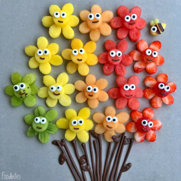 Colorful flower-shaped fruit bites with candy eyes are arranged in rows like a bouquet with chocolate stems. A small bee made from fruit rests near the top right flower, making these playful treats perfect for fun and healthy food recipes.