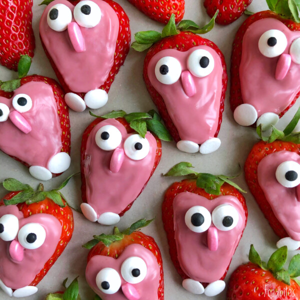 Strawberries dipped in pink icing are decorated with candy eyes, noses, and small white feet to look like cute cartoon faces—adorable bites perfect for fun food recipes or creative dishes, arranged on a light surface.