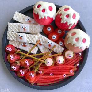 A black plate holds apples with white chocolate drips and candy eyes, wafer cookies and strawberries with candy eyes, pretzel sticks, mini mozzarella balls with olives, grape tomatoes, and red candy strips—fun bites perfect for health-conscious food recipes.