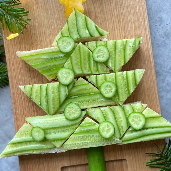 Triangle-shaped sandwiches topped with cucumber slices are arranged on a wooden board to resemble a Christmas tree, creating festive bites that add charm and health to your holiday food recipes.