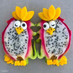 Two halved dragon fruits decorated to look like cartoon owls with mango slices for crests and feet, candy eyes, and small fruit pieces for beaks—these fun bites make creative and healthy dishes for food recipes. Arranged on a gray surface.