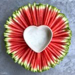 A round platter of neatly arranged watermelon sticks surrounds a heart-shaped bowl filled with white dipping sauce, creating one of those refreshing bites perfect for health-conscious food recipes. All set on a gray surface.