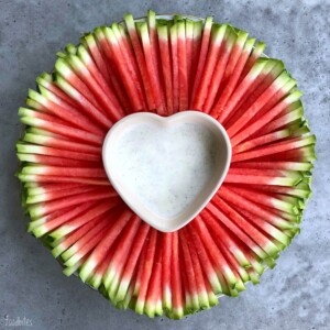 A round platter of neatly arranged watermelon sticks surrounds a heart-shaped bowl filled with white dipping sauce, creating one of those refreshing bites perfect for health-conscious food recipes. All set on a gray surface.