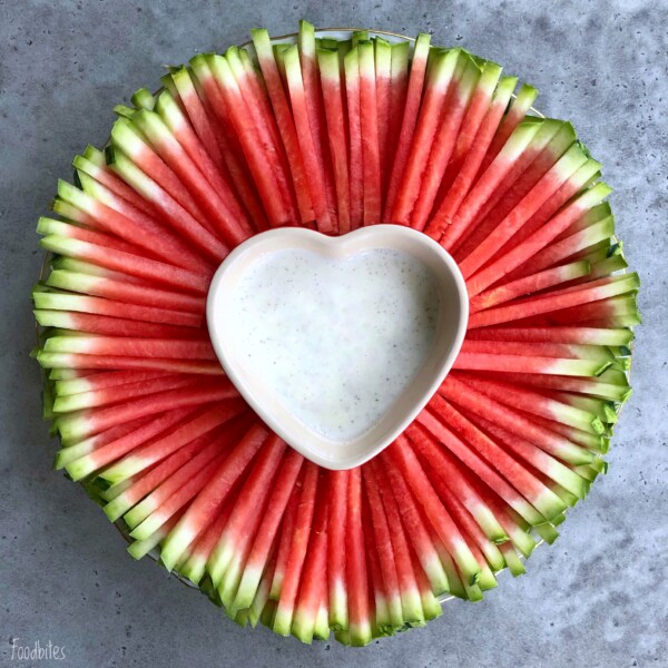 A round platter of neatly arranged watermelon sticks surrounds a heart-shaped bowl filled with white dipping sauce, creating one of those refreshing bites perfect for health-conscious food recipes. All set on a gray surface.
