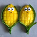 Two mango halves carved like corn on the cob, featuring grid patterns, green leaves, and decorated with candy eyes and smiles, make for fun bites and creative food recipes, placed side by side on a gray surface.