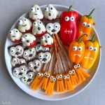 A plate of Halloween-themed bites: strawberries and cookies decorated as ghosts, pretzel sticks with cheese strips and candy eyes as brooms, and colorful bell peppers with faces—a fun way to enjoy creative food recipes.