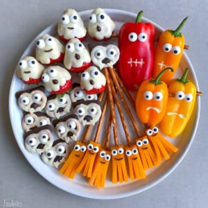 A plate of Halloween-themed bites: strawberries and cookies decorated as ghosts, pretzel sticks with cheese strips and candy eyes as brooms, and colorful bell peppers with faces—a fun way to enjoy creative food recipes.