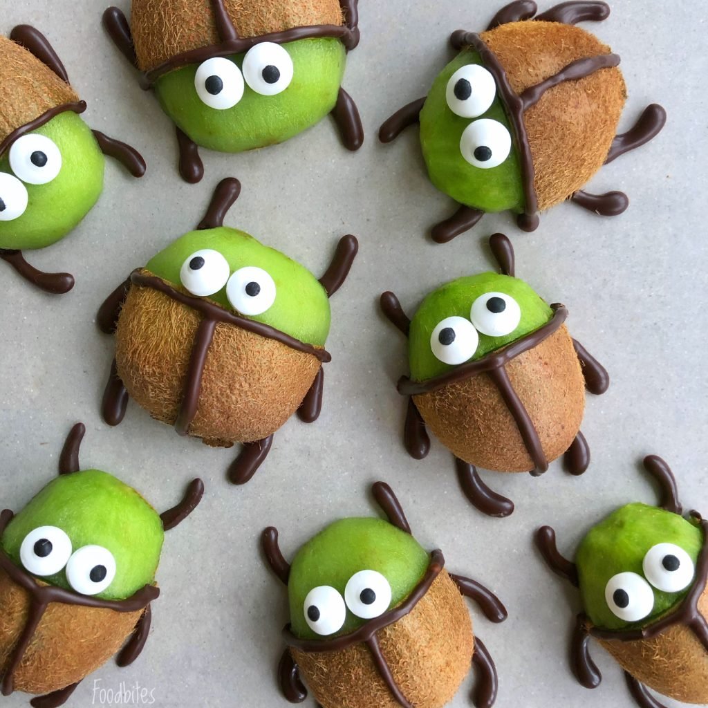 Kiwi halves are decorated with chocolate lines and candy eyes to look like cartoon beetles. They are arranged on a light grey surface.