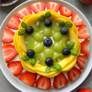 A plate topped with neatly arranged fresh fruit forms a colorful circular pattern—like a vibrant fruit pizza—with rings of sliced strawberries, mango slices, kiwi rounds, green grapes, blueberries, and basil leaves in the center.