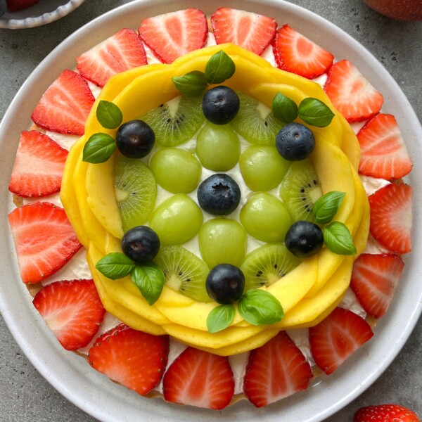 A plate topped with neatly arranged fresh fruit forms a colorful circular pattern—like a vibrant fruit pizza—with rings of sliced strawberries, mango slices, kiwi rounds, green grapes, blueberries, and basil leaves in the center.