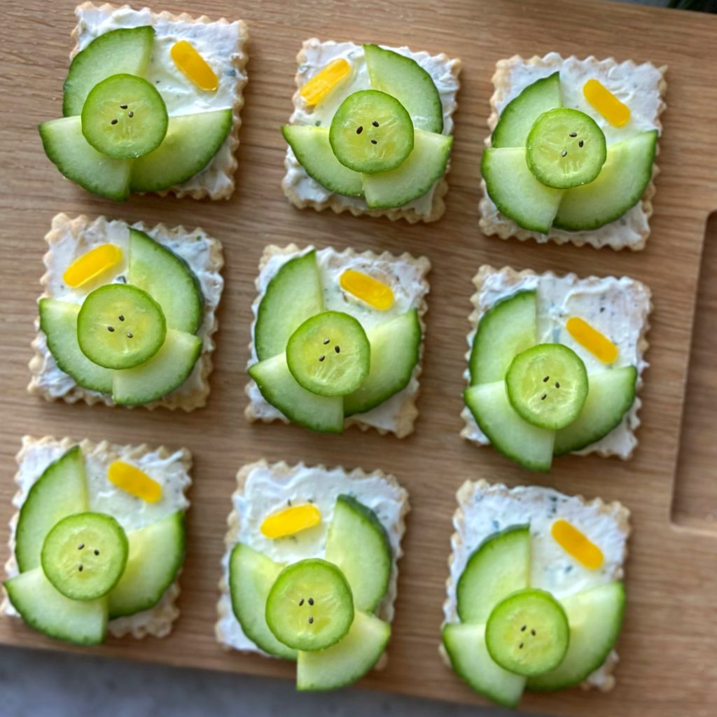 Closeup of nine crackers topped with a creamy spread, cucumber slices arranged as angels with small yellow pepper halos. They are displayed neatly on a wooden board.