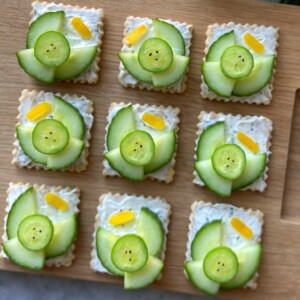 Nine crackers topped with a creamy spread, cucumber slices arranged in a flower pattern, and a small yellow garnish are displayed neatly on a wooden board—perfect for health-focused bites or creative food recipes.