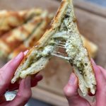 A close-up of hands pulling apart a grilled cheese sandwich, revealing gooey melted cheese and green pesto inside—delicious bites perfect for adding to your favorite food recipes, with more sandwich pieces blurred in the background.