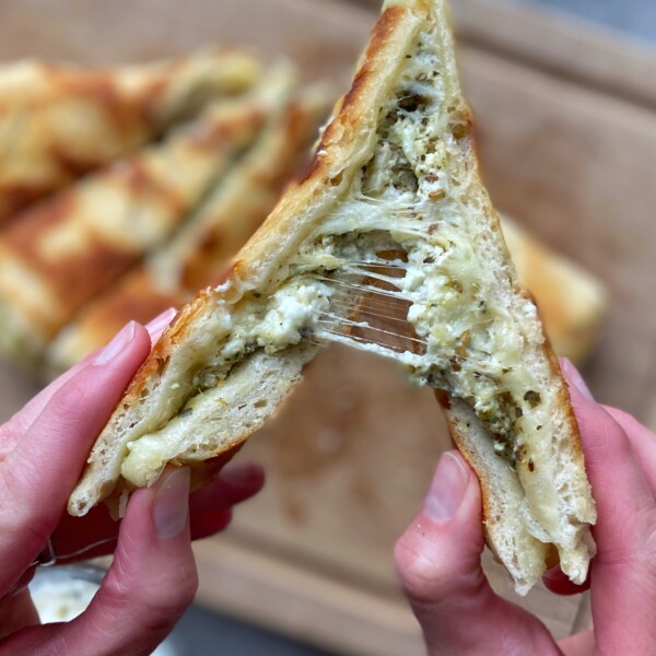 A close-up of hands pulling apart a grilled cheese sandwich, revealing gooey melted cheese and green pesto inside—delicious bites perfect for adding to your favorite food recipes, with more sandwich pieces blurred in the background.
