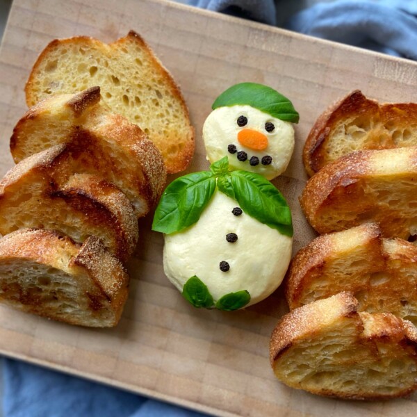 A cheese ball shaped like a snowman, decorated with basil leaves, black peppercorns, and a carrot nose, becomes the star of festive food recipes and dishes, placed on a wooden board surrounded by toasted bread slices.