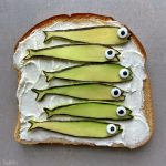 A slice of bread covered with white spread, topped with six avocado slices shaped like fish with candy eyes, arranged in a row to resemble fish swimming—perfect for creative bites and fun food recipes.