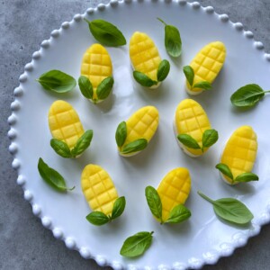 Sliced mango pieces shaped like mangoes with crosshatch patterns, arranged on a white plate with green basil leaves to resemble stems—perfect for healthy bites or creative food recipes.