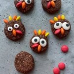 Chocolate cookies decorated with candy eyes and colorful candy pieces to look like cartoon turkeys are arranged on a gray surface, making these festive bites a fun addition to your holiday food recipes. Extra candies are scattered around for a playful touch.