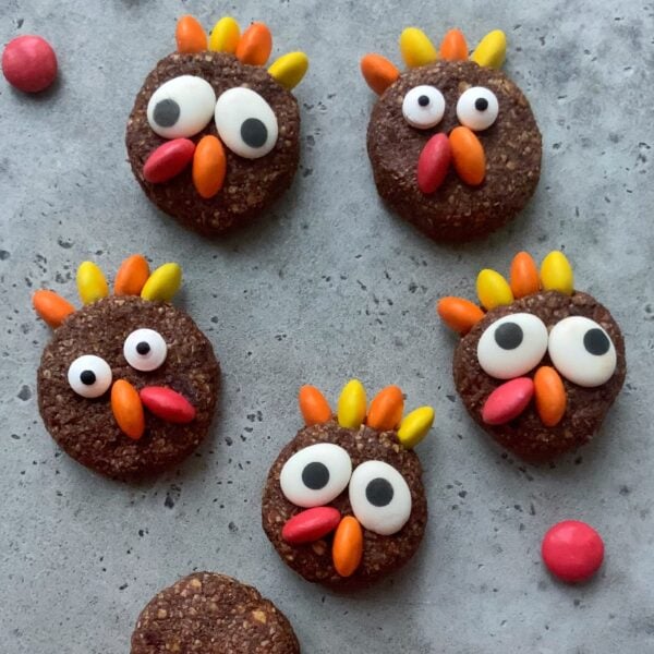 Chocolate protein balls decorated with candy eyes and colorful candy pieces to look like cartoon turkeys are arranged on a gray surface.