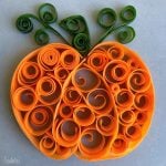An orange pumpkin shape made from curled and rolled strips of orange and green paper in a quilling art style, with green swirls at the top representing leaves and vines—perfect inspiration for health-themed food recipes or festive dishes.