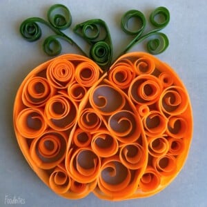 An orange pumpkin shape made from curled and rolled strips of orange and green paper in a quilling art style, with green swirls at the top representing leaves and vines—perfect inspiration for health-themed food recipes or festive dishes.