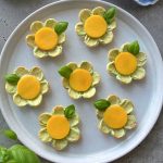 A plate of seven flower-shaped crackers topped with avocado appetizer spread, a yellow circular slice in the center, and a basil leaf garnish, arranged neatly on a round white plate.