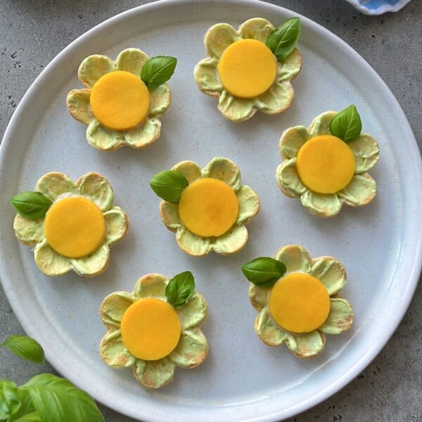 A plate of seven flower-shaped crackers topped with avocado appetizer spread, a yellow circular slice in the center, and a basil leaf garnish, arranged neatly on a round white plate.