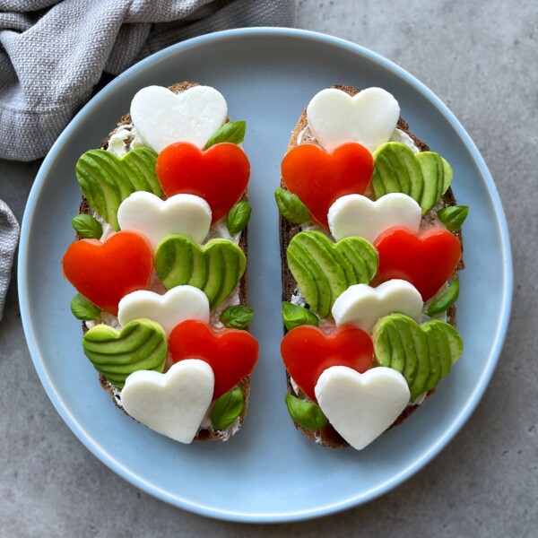 Two slices of bread topped with heart-shaped pieces of mozzarella, tomato, and avocado, arranged in a colorful pattern on a blue plate.