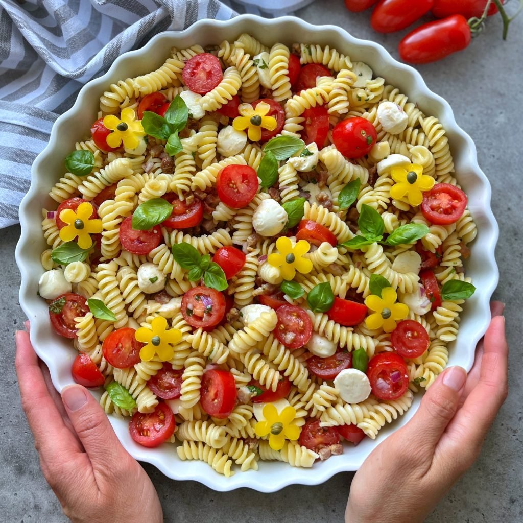 A person holds a round dish filled with fusilli pasta salad, cherry tomatoes, mozzarella balls, bacon, basil leaves, and yellow flower-shaped bites, on a gray surface with grape tomatoes and a striped tea towel nearby.