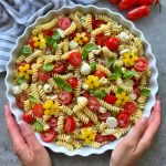 A person holds a round dish filled with healthy fusilli pasta salad, cherry tomatoes, mozzarella balls, basil leaves, and yellow flower-shaped bites, on a gray surface with grape tomatoes and a striped towel nearby.