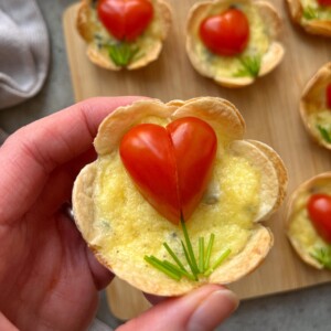 A hand holds a small Baked Egg Tortilla Bite with melted cheese, topped with a halved cherry tomato arranged as a heart and chive garnish. More tarts are visible on a wooden board in the background.