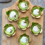Seven flower-shaped bites made of overlapping potato chips, sliced avocado, and a dollop of white sauce on a wooden board. Some are garnished with green herbs or basil leaves—perfect for health-conscious food recipes and appetizers.
