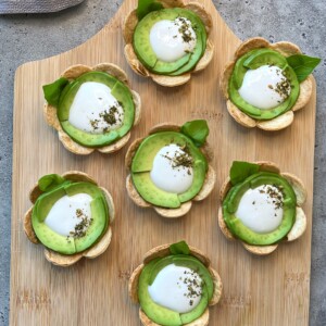 Seven flower-shaped bites made of overlapping potato chips, sliced avocado, and a dollop of white sauce on a wooden board. Some are garnished with green herbs or basil leaves—perfect for health-conscious food recipes and appetizers.
