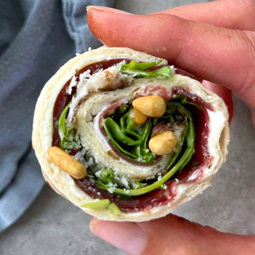 A hand holds a pinwheel-shaped wrap filled with leafy greens, cream cheese, fruit preserves, and a few nuts, making for one of those tasty health bites rolled in a tortilla.
