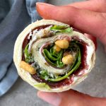 A hand holds a pinwheel-shaped wrap filled with leafy greens, cream cheese, fruit preserves, and a few nuts, making for one of those tasty health bites rolled in a tortilla.