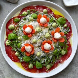 A vibrant Beef Carpaccio Salad features thin tomato slices, arugula, basil leaves, avocado cream dollops, grated cheese, and mozzarella balls with carrot flowers. All are drizzled with olive oil on a pristine white plate.