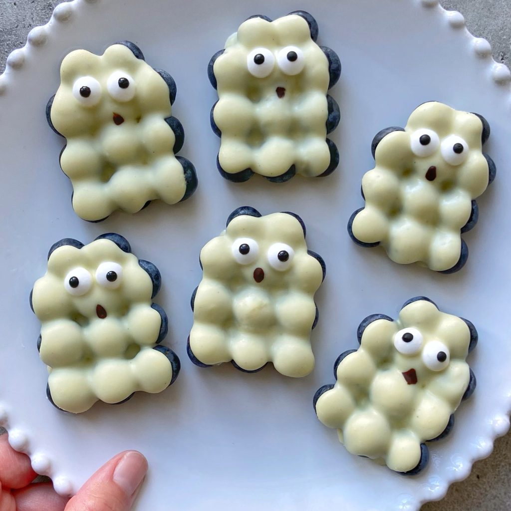 Six white chocolate blueberry bites shaped like bubbly ghosts, each with cute, surprised facial expressions and wide eyes, are arranged on a scalloped-edge white plate. A hand holds the edge the plate.