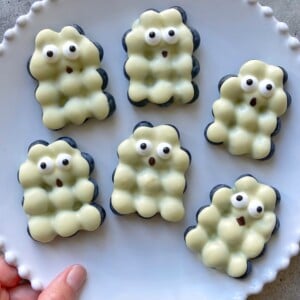 Six white chocolate blueberry bites shaped like bubbly ghosts, each with cute, surprised facial expressions and wide eyes, are arranged on a scalloped-edge white plate. A hand holds the edge the plate.