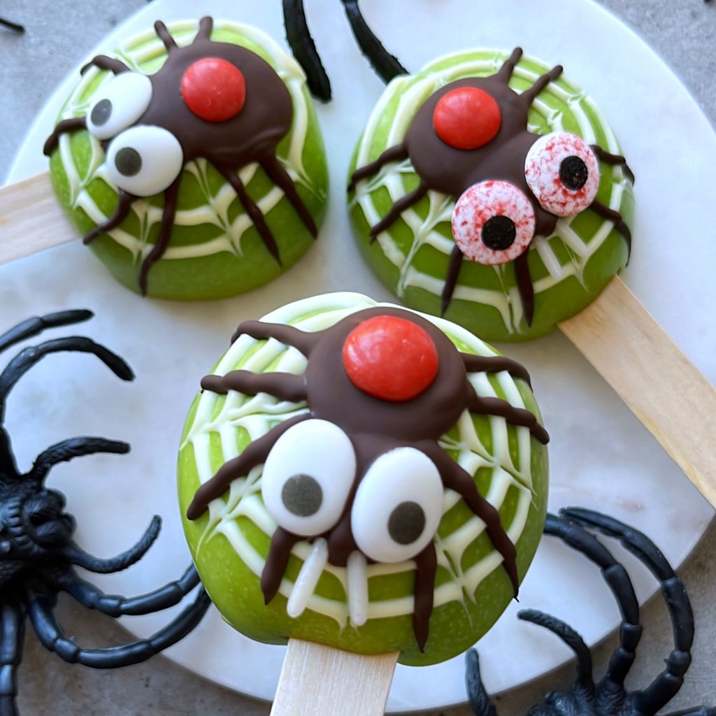 Green apple slices on sticks decorated with chocolate, candy eyes, and red candies to look like spiders on webs. The apples are arranged on a plate surrounded by plastic toy spiders.
