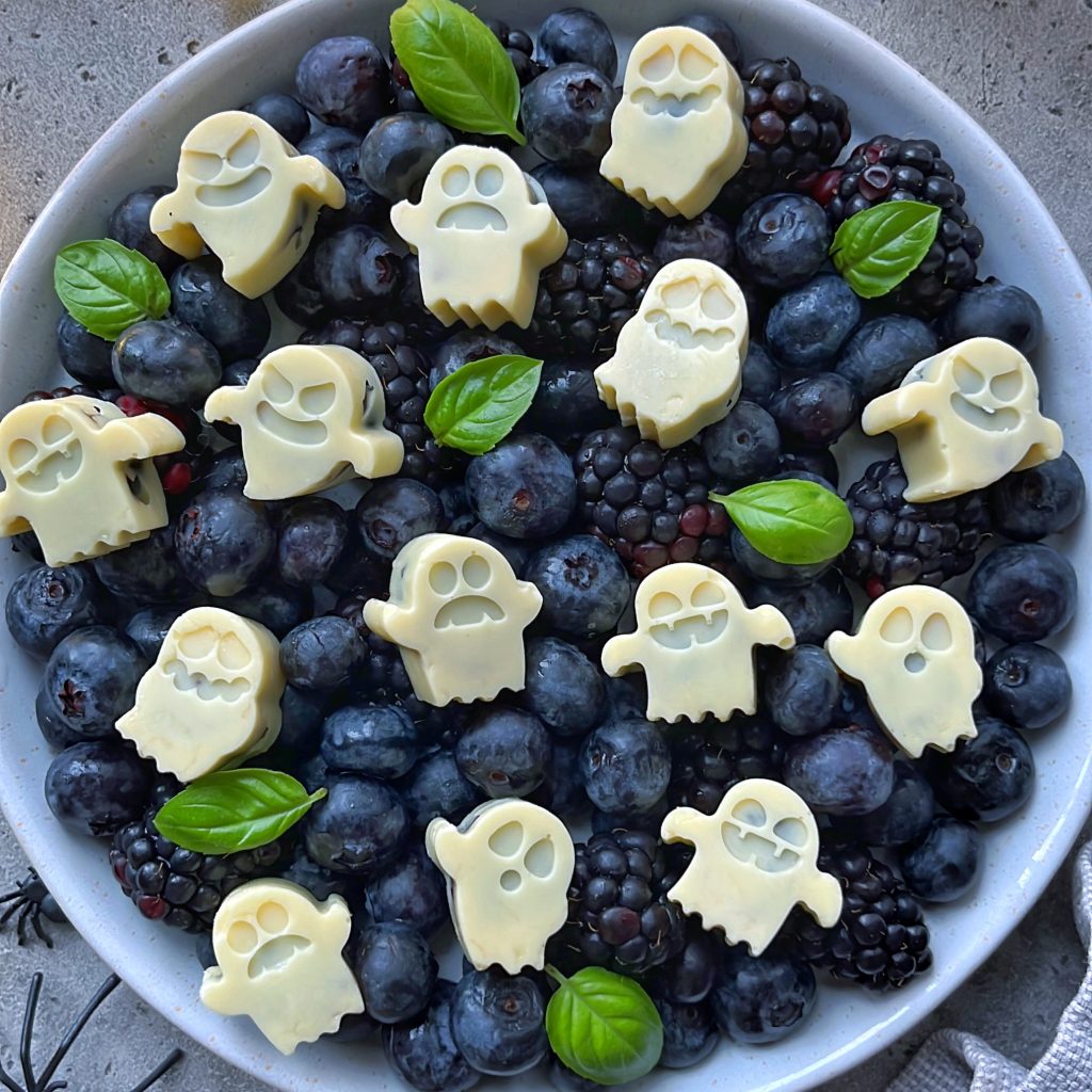 A bowl filled with blackberries and blueberries, topped with white chocolate pieces shaped like ghosts—Blueberry Ghost Bites—and garnished with fresh green basil leaves.