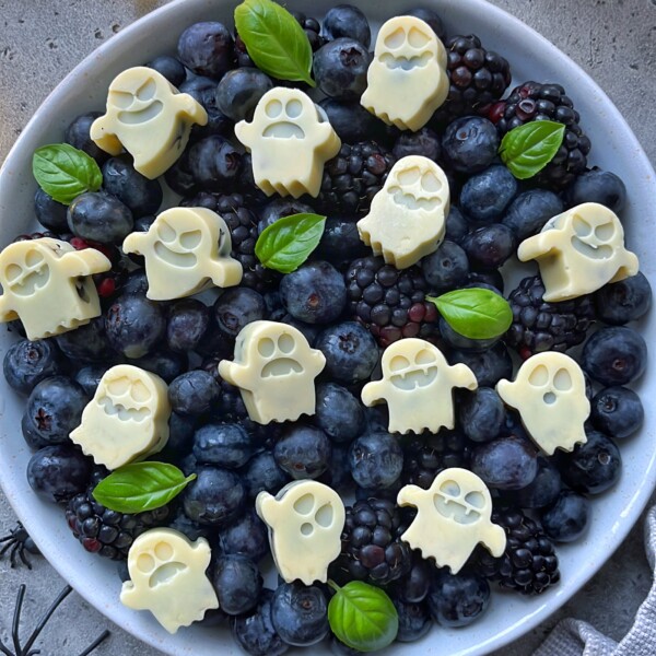 A bowl filled with blackberries and blueberries, topped with white chocolate pieces shaped like ghosts—Blueberry Ghost Bites—and garnished with fresh green basil leaves.