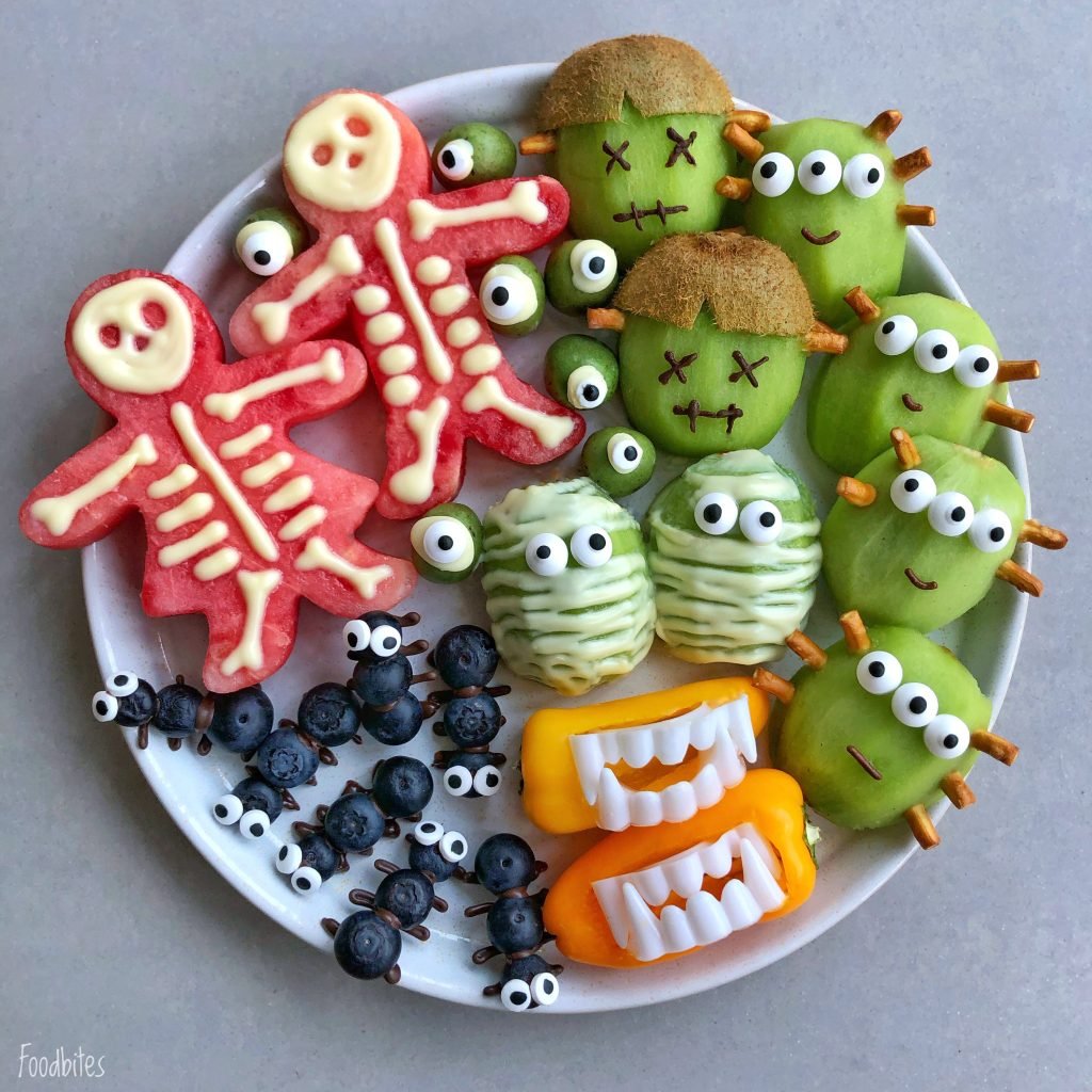 A plate of creatively arranged fruits and veggies makes healthy Halloween bites: watermelon skeletons, kiwi monsters, green apple mummies, blueberry spiders, and bell pepper mouths with teeth.